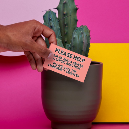 Person holding a severe allergic reaction alert card next to a potted cactus on a pink and yellow background