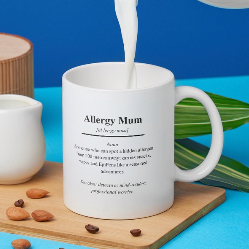 White mug with 'Allergy Mum' definition text, milk being poured into it on a blue background.